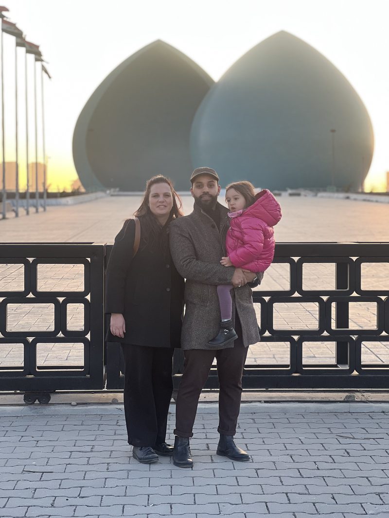 Family of three tourists posing in front of the Martyr Monument at sunset with the turquoise domes visible in golden light, Baghdad