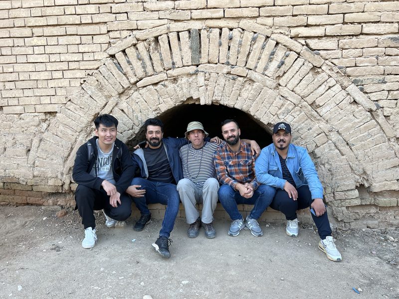 Group of four tourists sitting and standing under an ancient brick arched doorway at the ruins of Babylon, Iraq