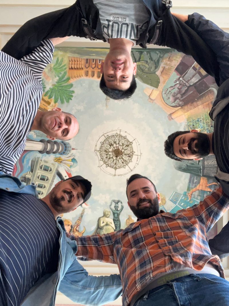 Group of four tourists and guides looking up at a colorful painted ceiling mural depicting Iraqi landmarks in Saddam Hussein Palace
