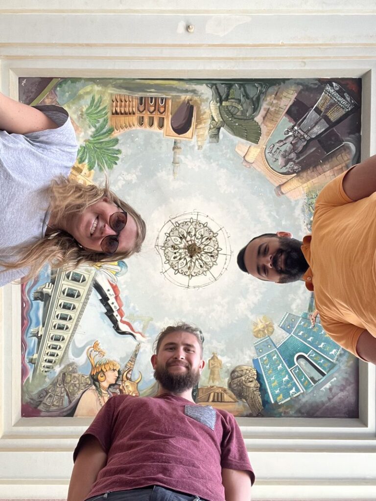 Two tourists looking up and posing around a colorful painted ceiling mural depicting famous Iraqi Saddam Hussein Palace
