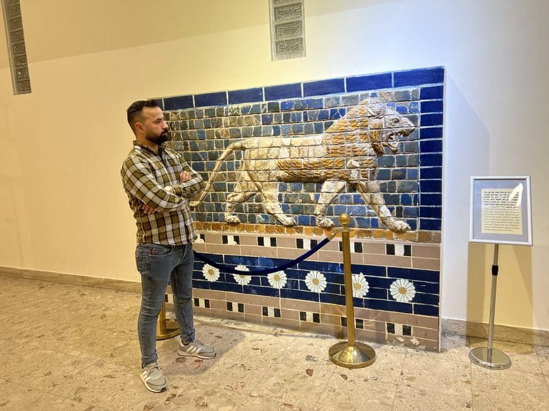 Ali Babylon tour guide standing beside a reconstructed Ishtar Gate lion mosaic panel at a museum in Iraq