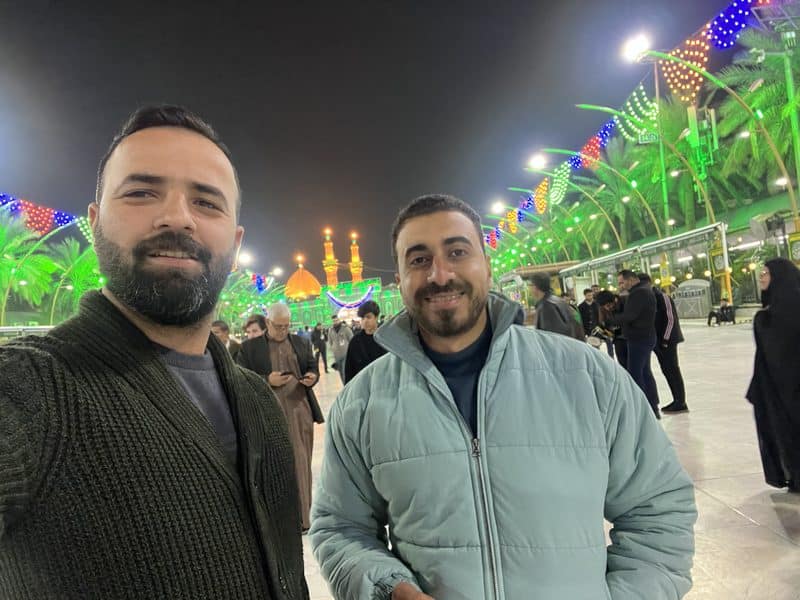 Ali Babylon tour guide taking a selfie at night with the illuminated Imam Hussein Shrine glowing green in the background in Karbala, Iraq