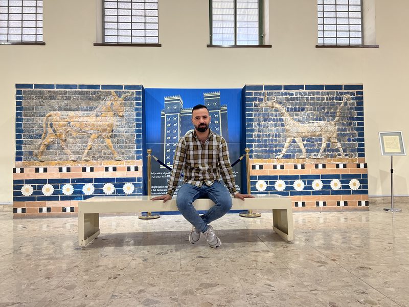 Ali Babylon tour guide sitting in front of a large reconstructed Ishtar Gate mosaic with dragon and bull reliefs at the Iraq Museum in Baghdad