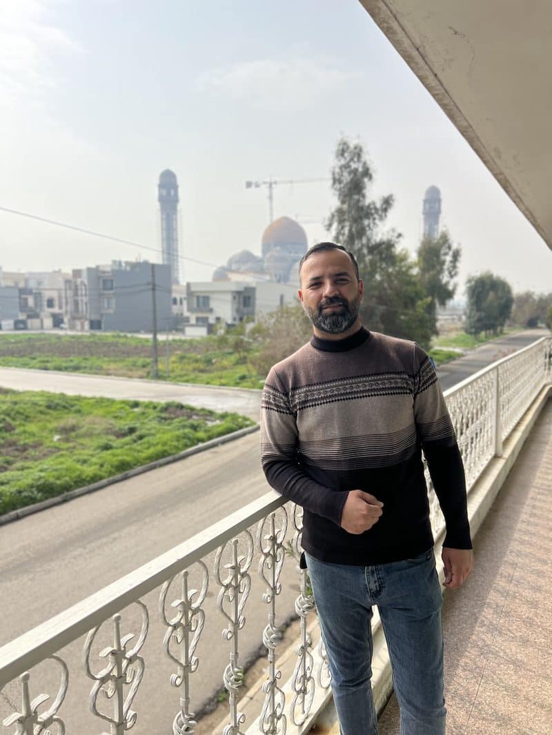 Ali Babylon tour guide standing on a balcony with a mosque and golden dome in the background in Hillah, Iraq