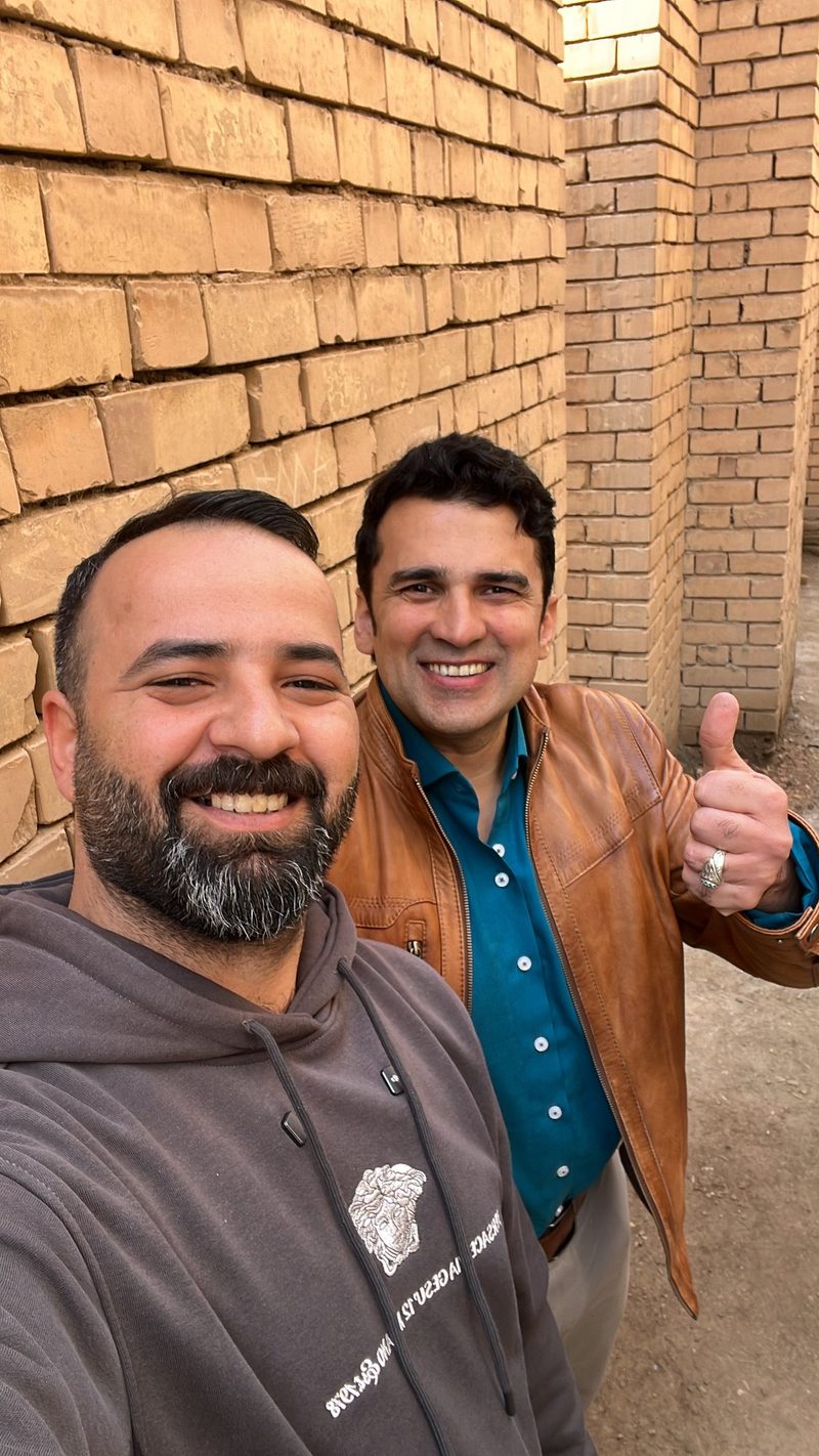 Ali Babylon tour guide taking a selfie with a smiling guest giving thumbs up in front of ancient mud brick walls at an archaeological site in Iraq