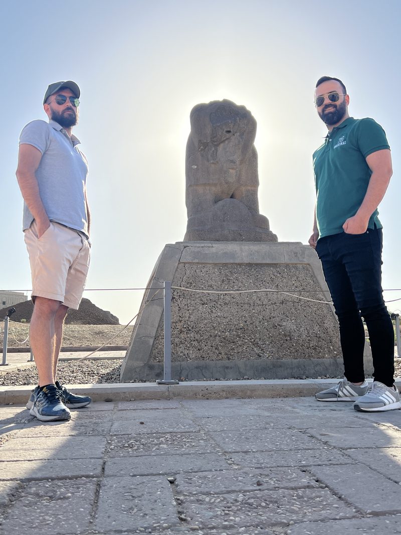 Two men standing on either side of the ancient Lion of Babylon stone statue backlit against a bright sky at the Babylon archaeological site
