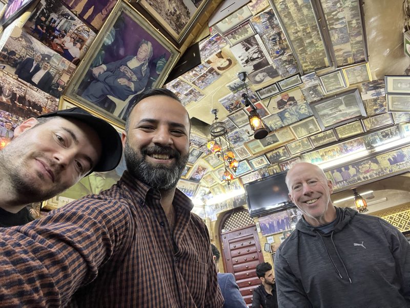 Ali Babylon tour guide taking a selfie with two guests inside a traditional Iraqi café decorated with vintage photos and memorabilia