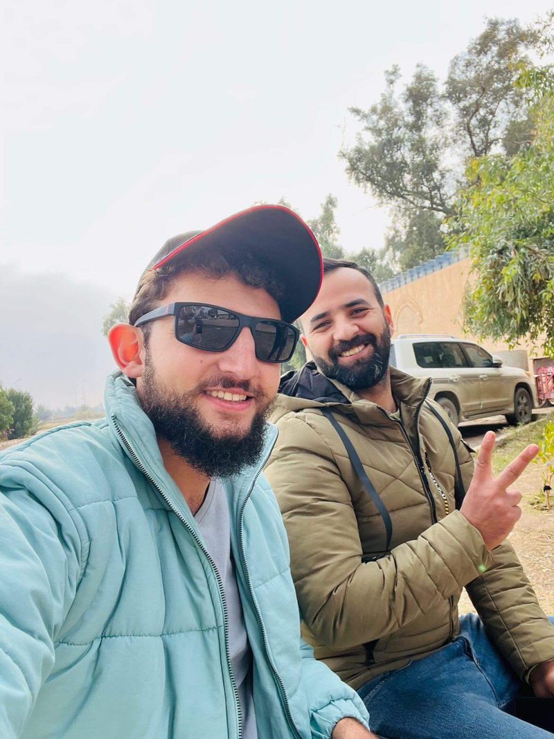 Two men taking a casual selfie outdoors near a tree in Iraq, one wearing sunglasses and giving a peace sign