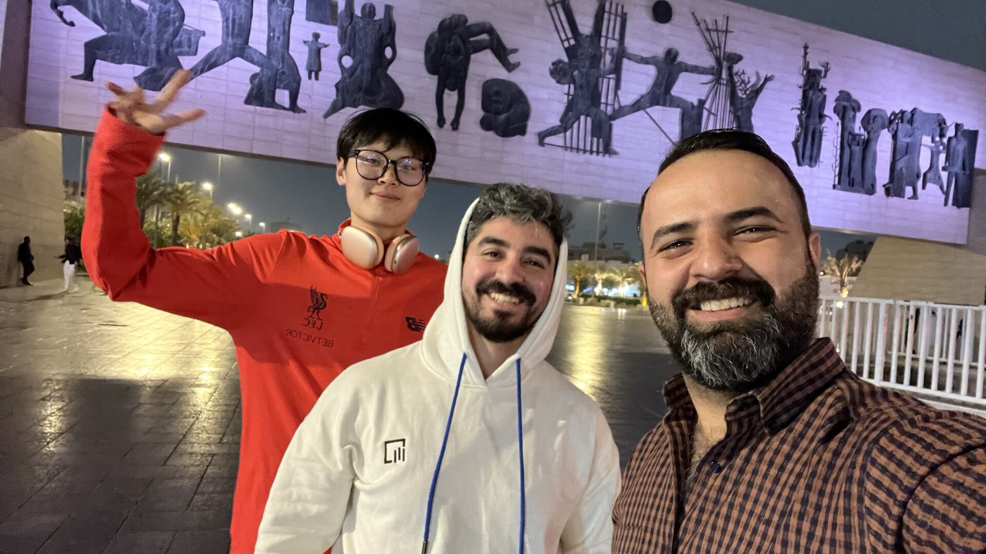 Two smiling travelers with Ali posing for a selfie at night in front of Baghdad's iconic Martyr Monument, its large bas-relief mural illuminated against the evening sky.