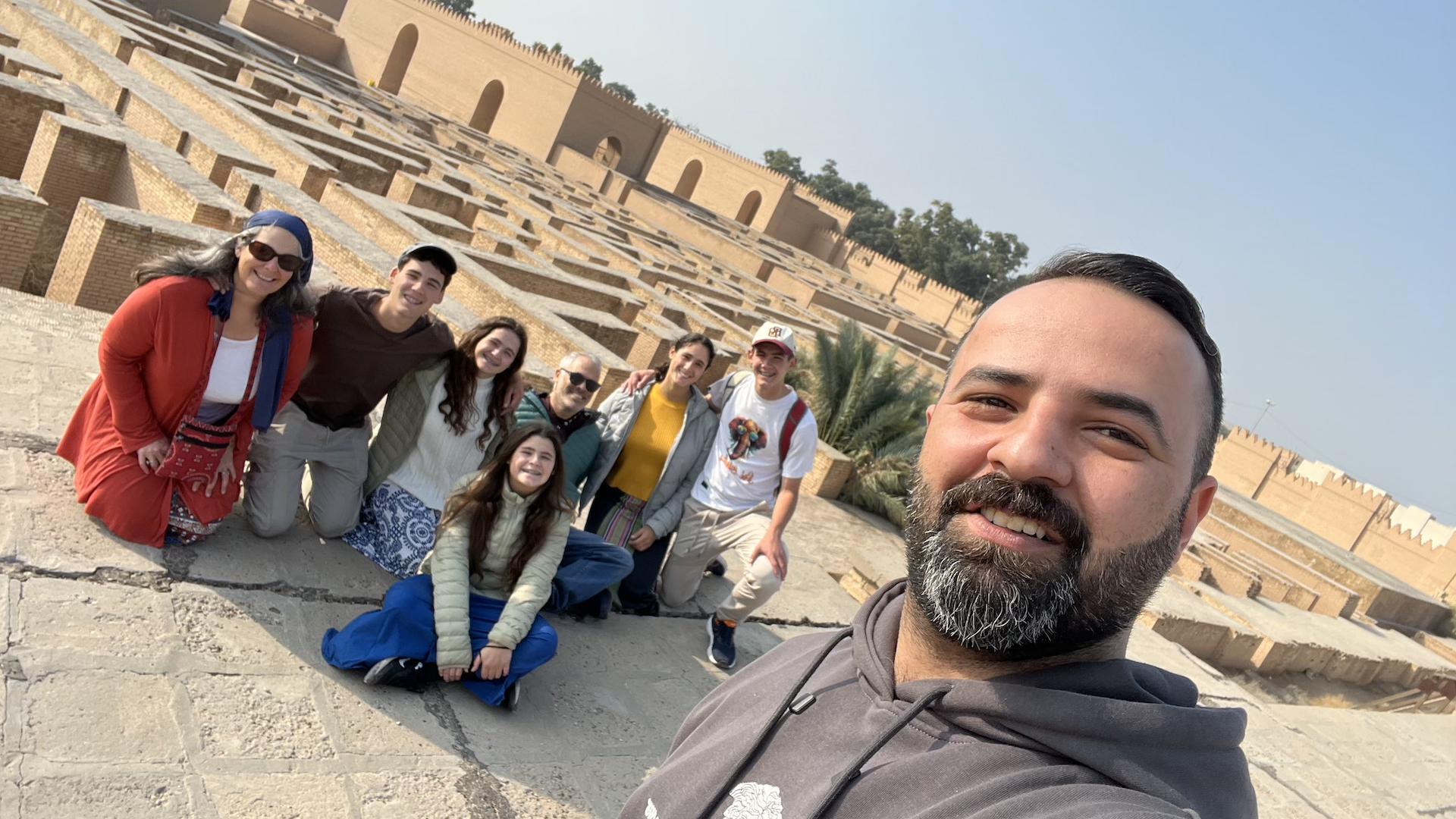 A tour group of seven people posing for a selfie on the ancient stone terraces of the ruins of Babylon, Iraq, with reconstructed mud-brick walls and palm trees in the background