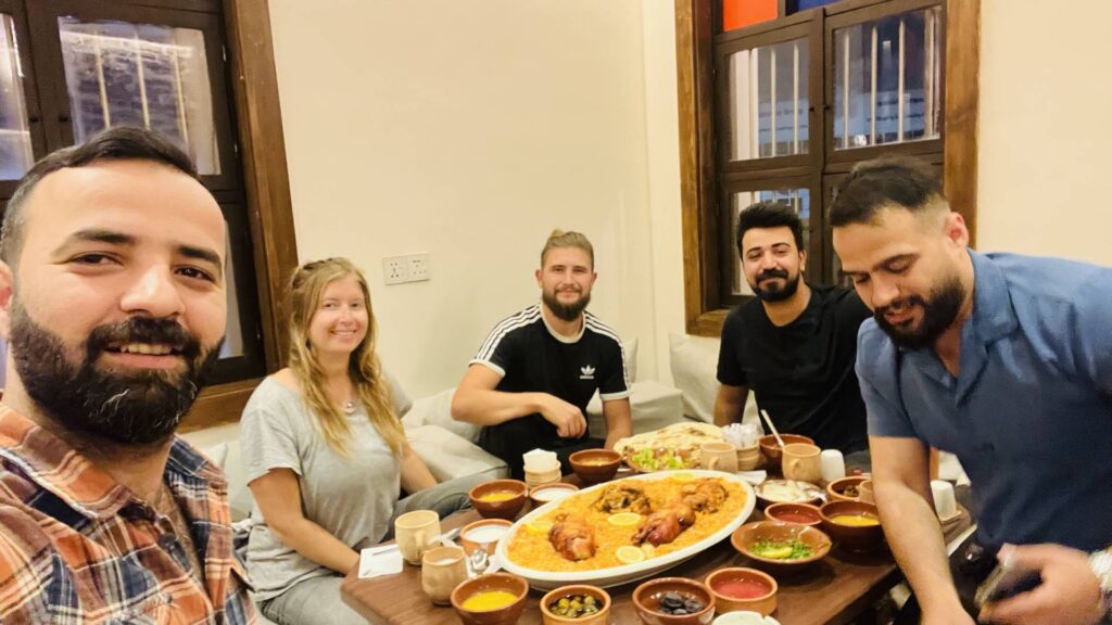 A group of five travelers sharing a traditional Iraqi feast with a large platter of rice and chicken surrounded by clay bowls of mezze and sides at a local restaurant