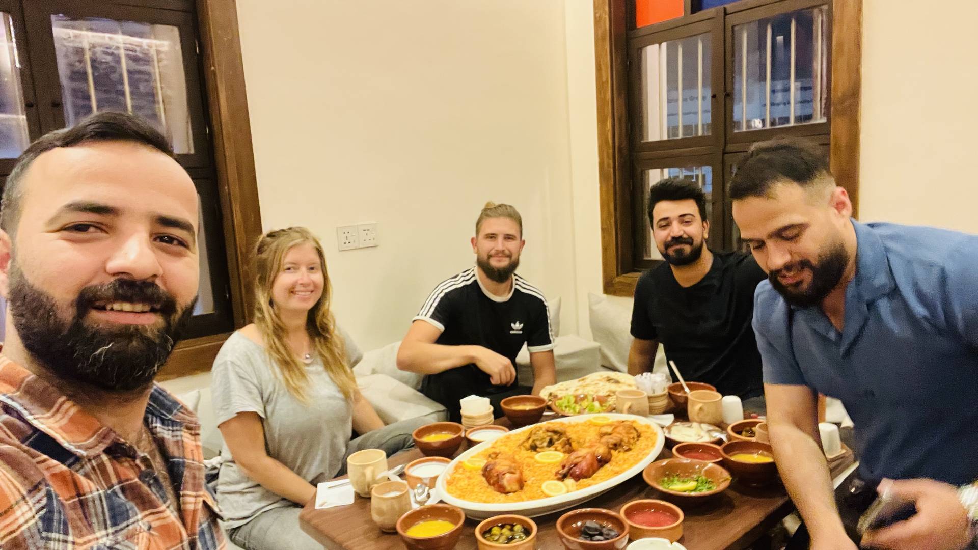 A group of five travelers sharing a traditional Iraqi feast with a large platter of rice and chicken surrounded by clay bowls of mezze and sides at a local restaurant