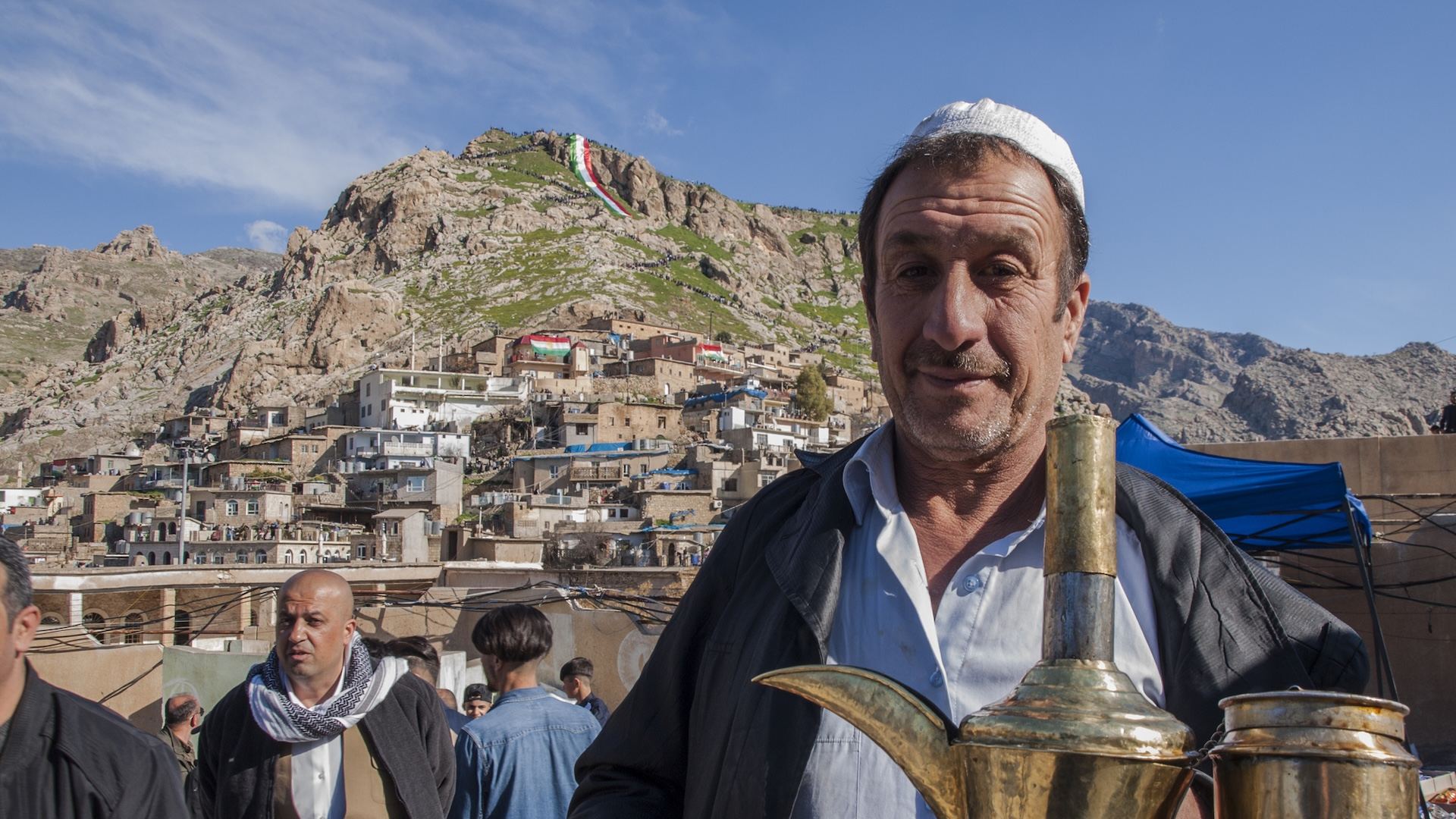 Kurdish tea vendor holding traditional brass dallah coffee pots at a busy market in Akre, with the historic hillside town and Kurdistan flag in the background