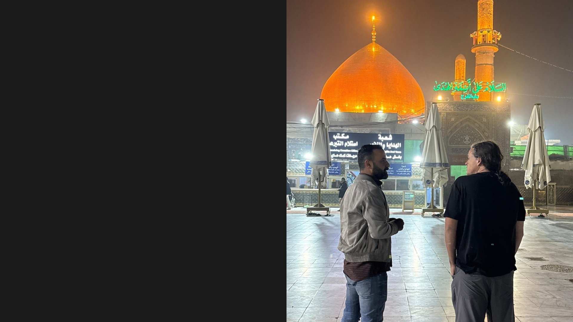 Two men standing in a courtyard at night in front of the illuminated golden dome and minarets of the Imam Ali Shrine in Karbala, Iraq, with Arabic signage visible in the background