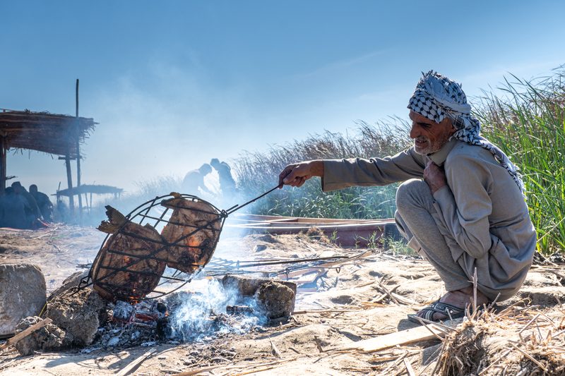 Marsh Arab man preparing traditional masgouf grilled fish over a campfire in southern Iraq
