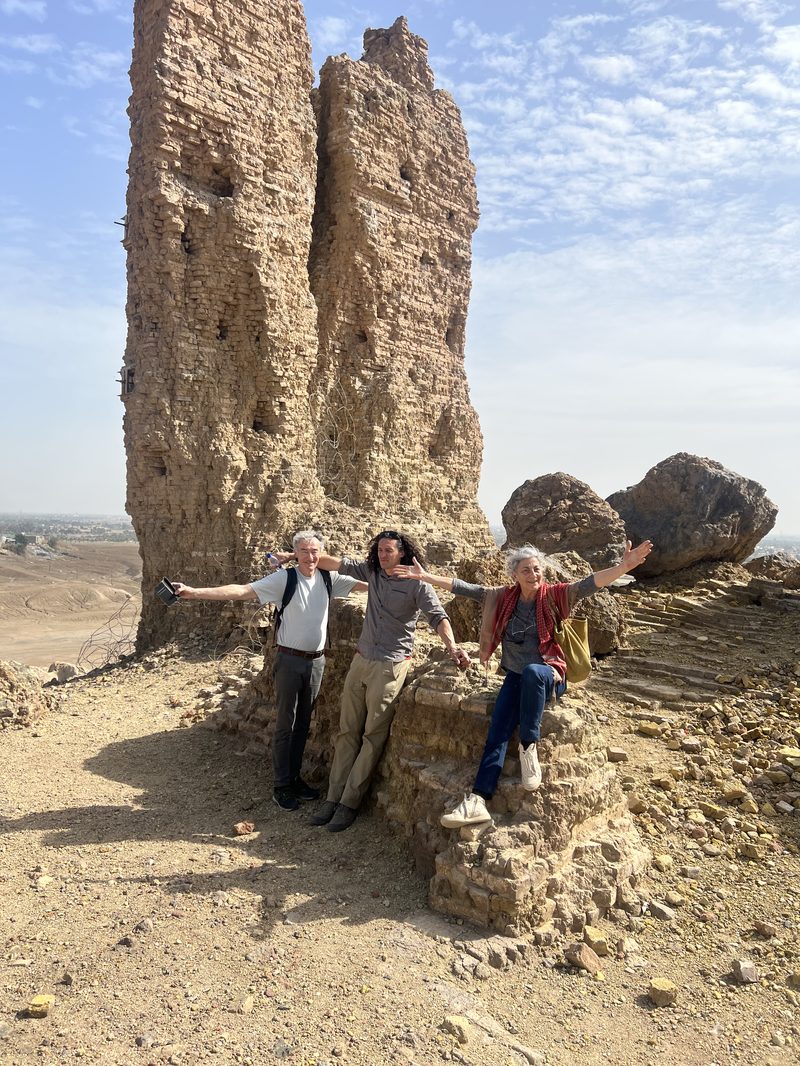 Three tourists with arms outstretched celebrating in front of tall ancient mud brick tower ruins at Nineveh near Mosul, Iraq